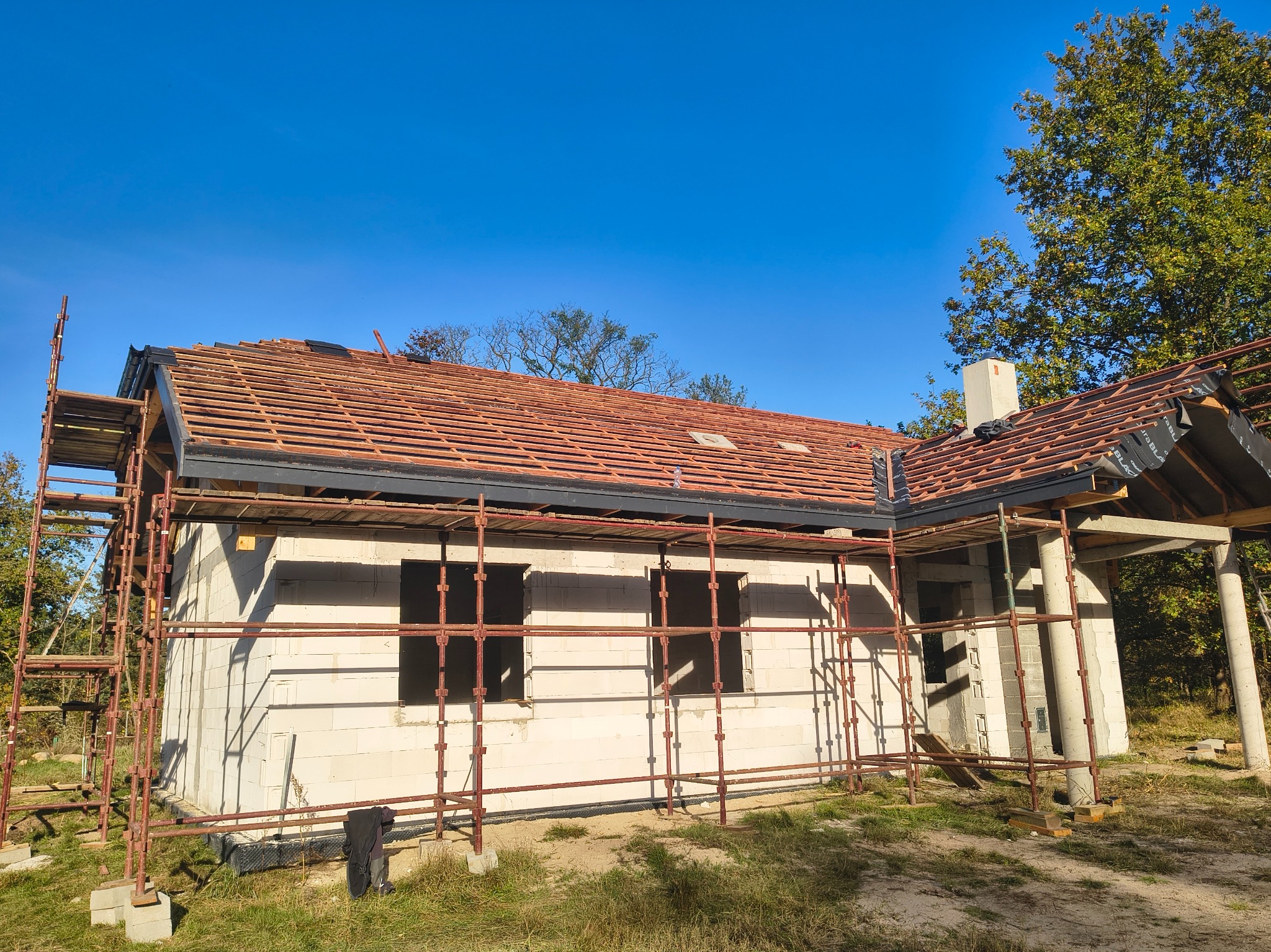 Wznoszony dom z białego pustaka, z częściowo ułożoną czerwoną dachówką na łatach. Widoczne rusztowania i fragmenty drzew na tle błękitnego nieba.