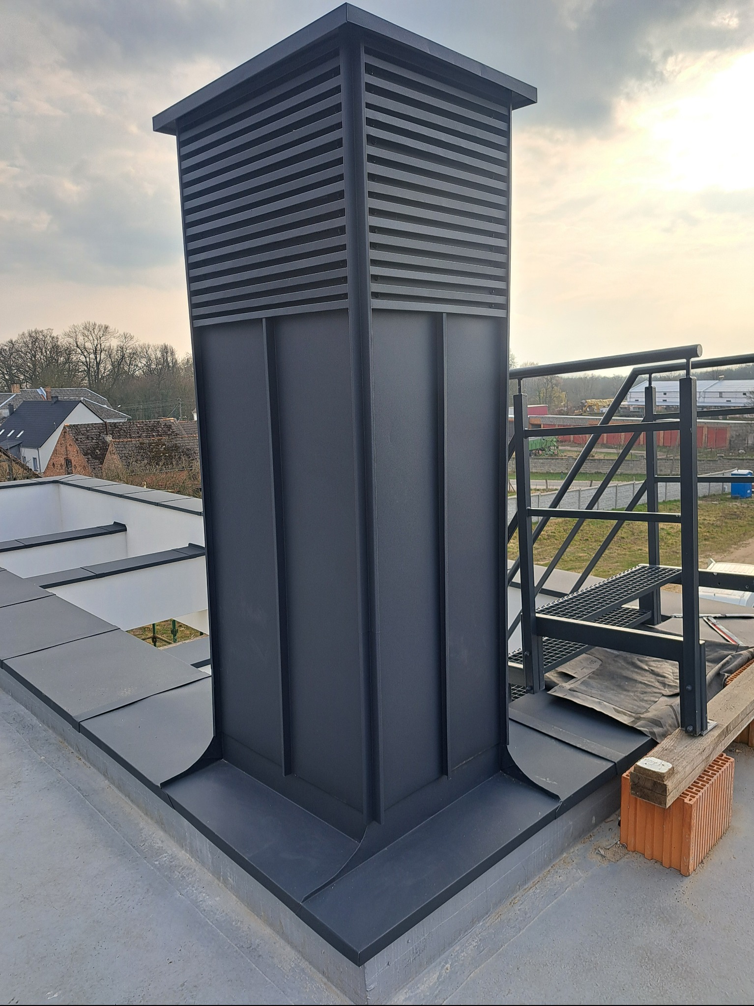 Nowoczesny komin wentylacyjny z blachy na płaskim dachu, z widoczną drabinką i elementami wykończeniowymi. Precyzyjne obróbki blacharskie.