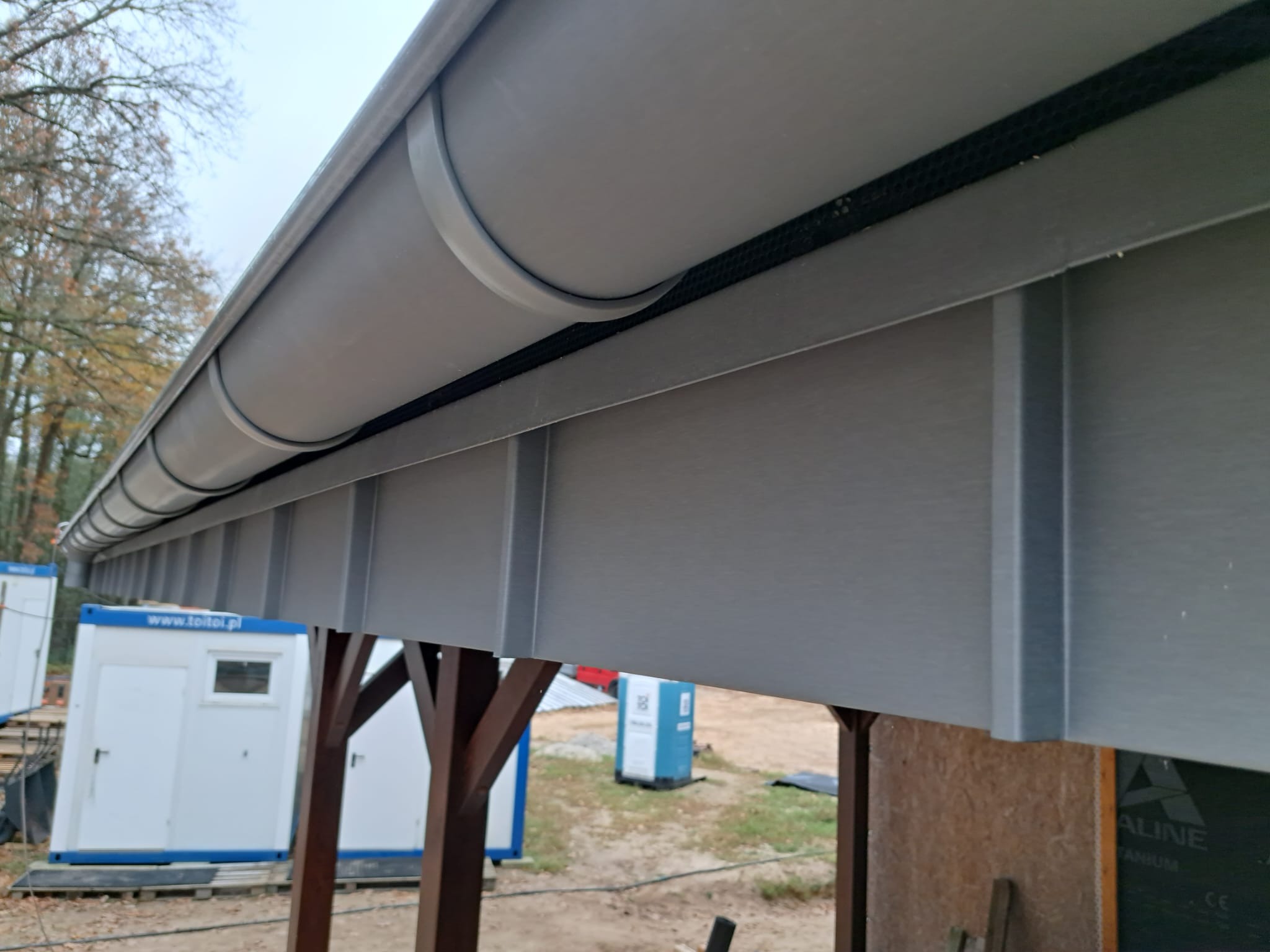 Szara rynna dachowa z elementami mocującymi, widoczna z bliska pod kątem. W tle kontenery budowlane i drzewa.