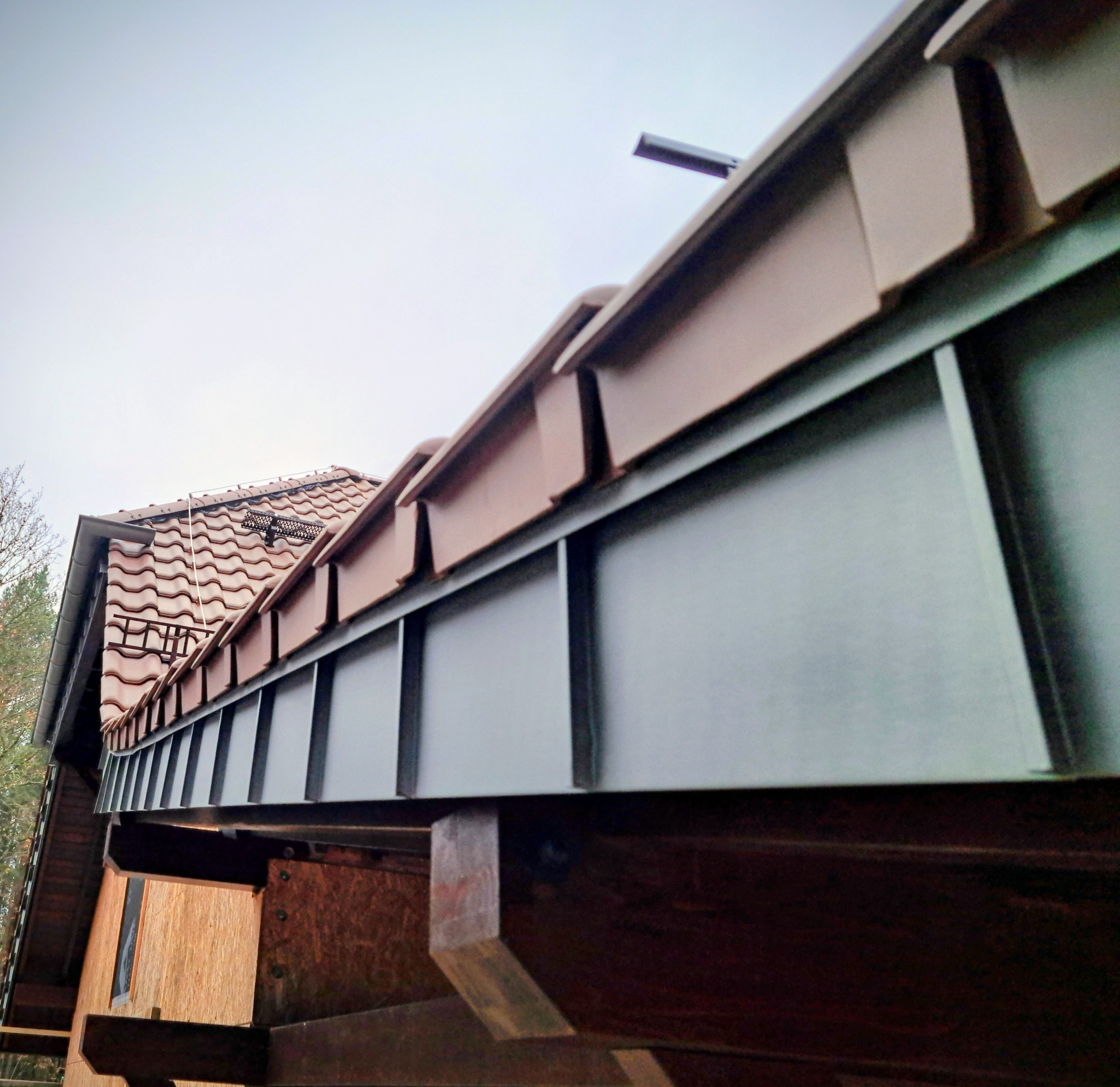 Szczegółowe ujęcie nowej, metalowej rynny w kolorze grafitowym, zamontowanej na drewnianej konstrukcji dachu z brązową dachówką ceramiczną w tle.