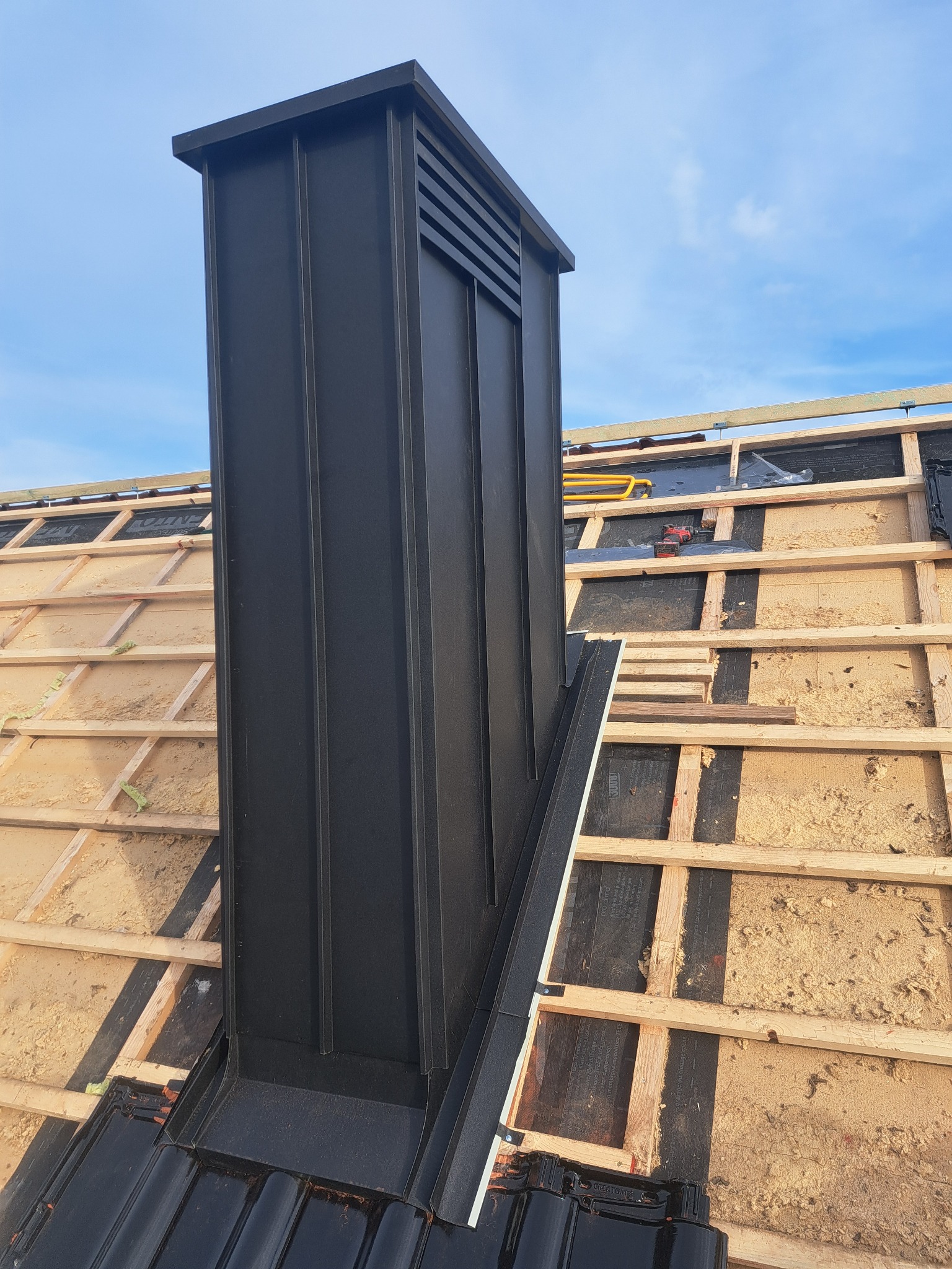Czarny komin wentylacyjny z blachy na dachu w trakcie budowy. Widoczne drewniane elementy konstrukcji dachu i fragmenty czarnej dachówki. Błękitne niebo w tle.