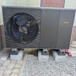 Zamontowana pompa ciepła Midea na betonowych podstawach, obok budynku z widoczną fakturą elewacji i fragmentem chodnika z kostki brukowej. Widoczny wentylator.