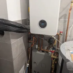 Nowoczesny, biały piec gazowy Bosch z podłączeniami miedzianymi na tle szarych płytek. Widoczny zbiornik i elementy instalacji. Estetyczny montaż w kotłowni.