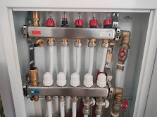 Rozdzielacz instalacji grzewczej z białymi rurami, mosiężnymi złączkami i czerwonymi zaworami w białej szafce. Widok z przodu na precyzyjnie połączone elementy.