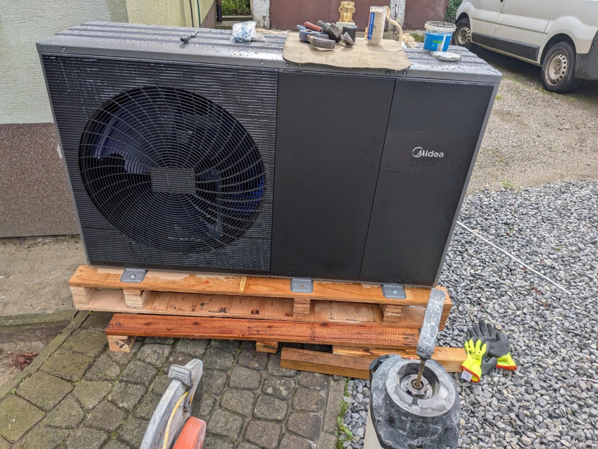 Pompa ciepła Midea na palecie przed montażem, widoczne narzędzia i rękawice robocze. Na obudowie widoczne krople deszczu. Obok zaparkowany biały samochód dostawczy.