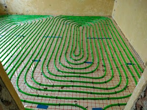 Zielone rury ogrzewania podłogowego ułożone spiralnie na czerwonej siatce w jasnym pomieszczeniu przed wylaniem posadzki. Widoczne fragmenty ścian.