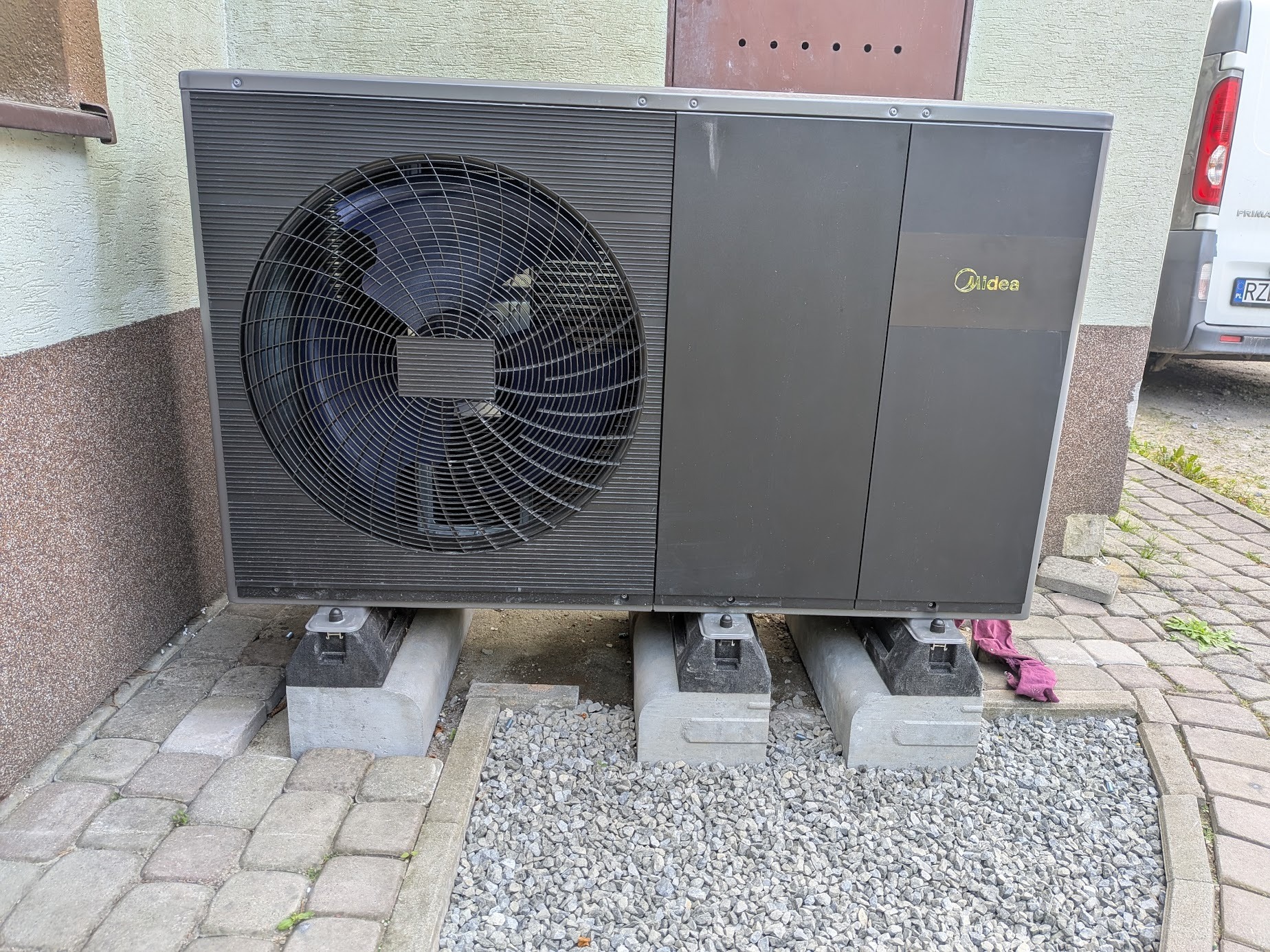 Zamontowana pompa ciepła Midea na betonowych podstawach, obok budynku z widoczną fakturą elewacji i fragmentem chodnika z kostki brukowej. Widoczny wentylator.