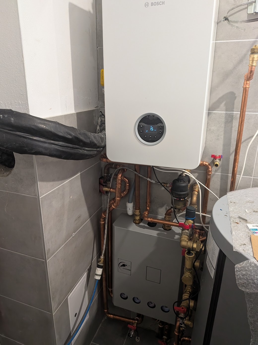 Nowoczesny, biały piec gazowy Bosch z podłączeniami miedzianymi na tle szarych płytek. Widoczny zbiornik i elementy instalacji. Estetyczny montaż w kotłowni.