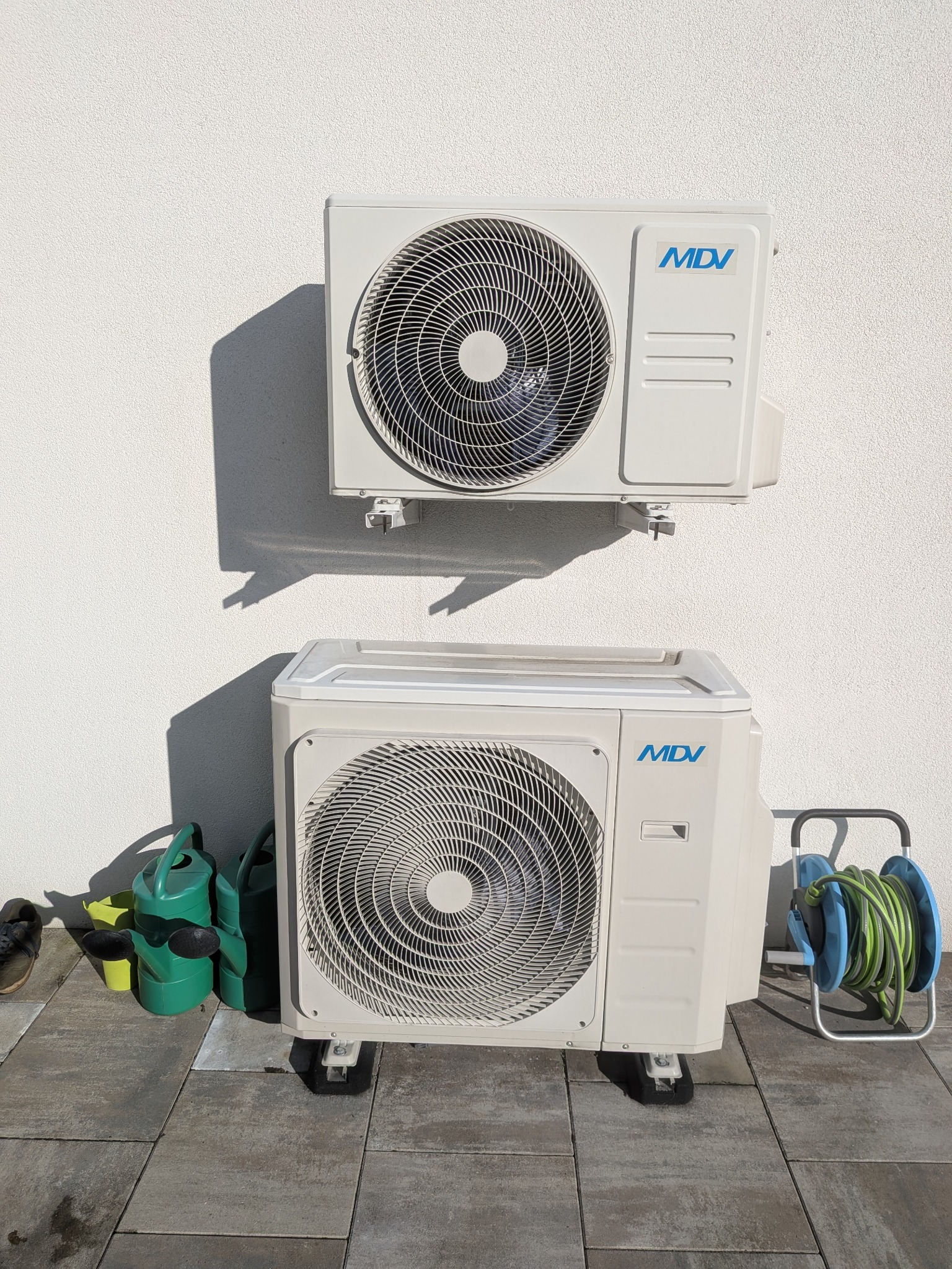 Dwie jednostki zewnętrzne klimatyzacji MDV zamontowane na białej ścianie. Obok dolnej jednostki widać zielone konewki i zwinięty wąż ogrodowy.