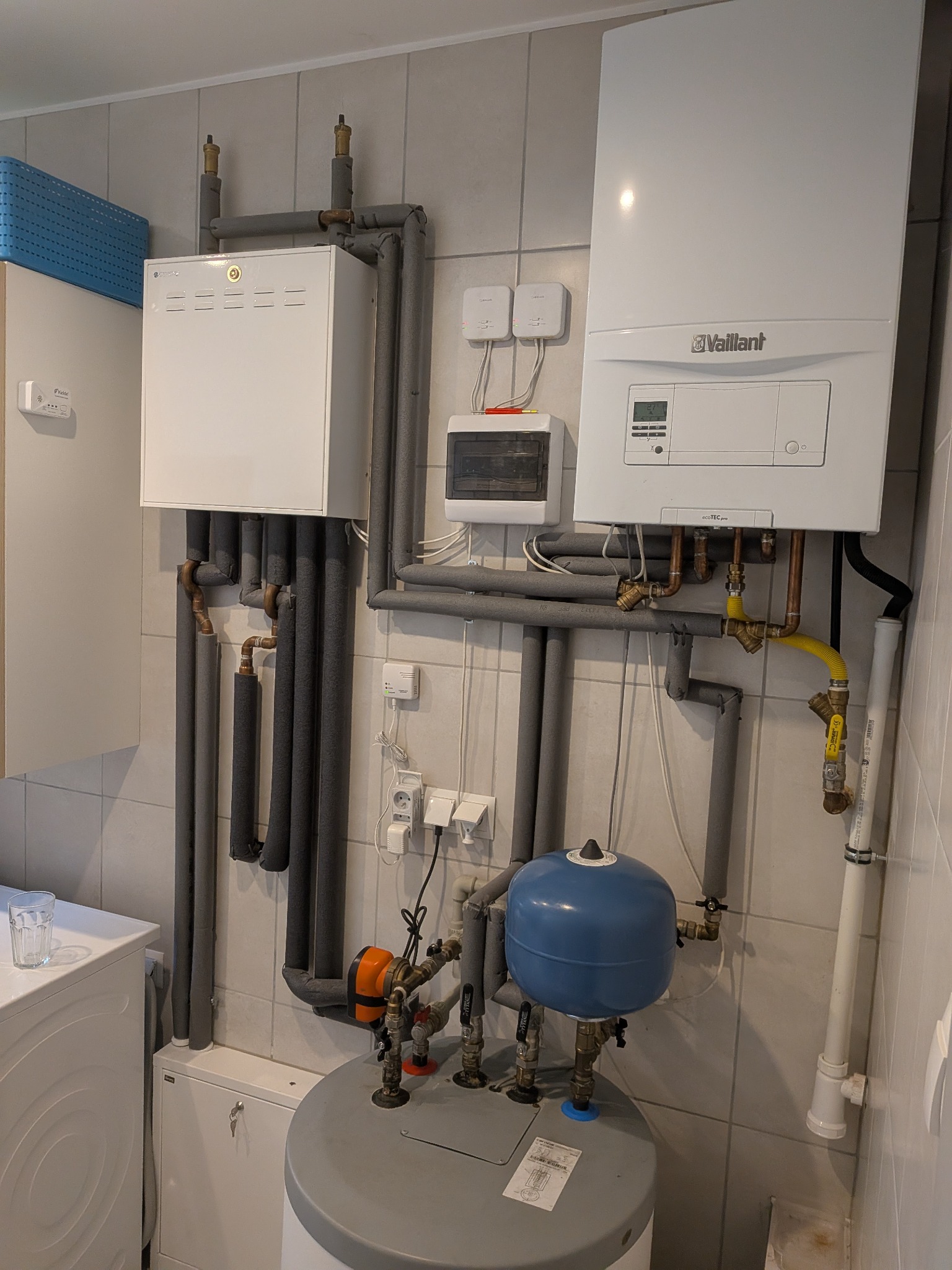 Biały piec gazowy Vaillant z szarymi rurami w izolacji, zbiornik wyrównawczy i instalacja elektryczna na tle płytek. Widok na kompletną instalację grzewczą w pomieszczeniu.