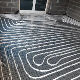 Instalacja ogrzewania podłogowego z rurkami grzewczymi ułożonymi na folii z nadrukowaną siatką, widoczne surowe ściany z bloczków betonowych i okno w tle.