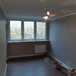 Pok&oacute;j po świeżym pomalowaniu na szaro, z widocznymi śladami po niedawnych pracach: kartonowe pudło, wiadro z farbą i szpachelka przy listwie przypodłogowej, oraz wentylator sufitowy.