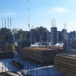 Wysoki kąt widzenia na plac budowy z widocznymi zbrojeniami, betonowymi kolumnami w szalunkach, stosami drewna i pracującymi robotnikami w oddali, pod błękitnym niebem.