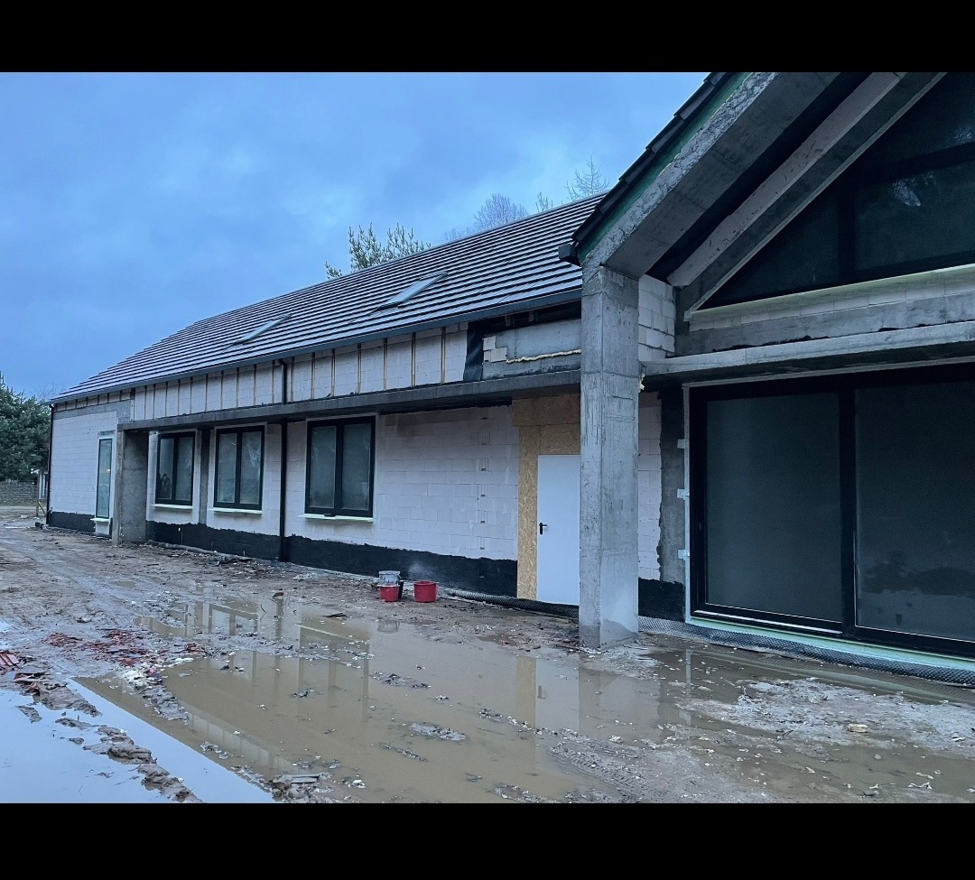 Surowy stan domu murowanego w Gdyni, z widocznymi oknami, drzwiami i częściowo wykończonym dachem, otoczony błotnistym terenem.