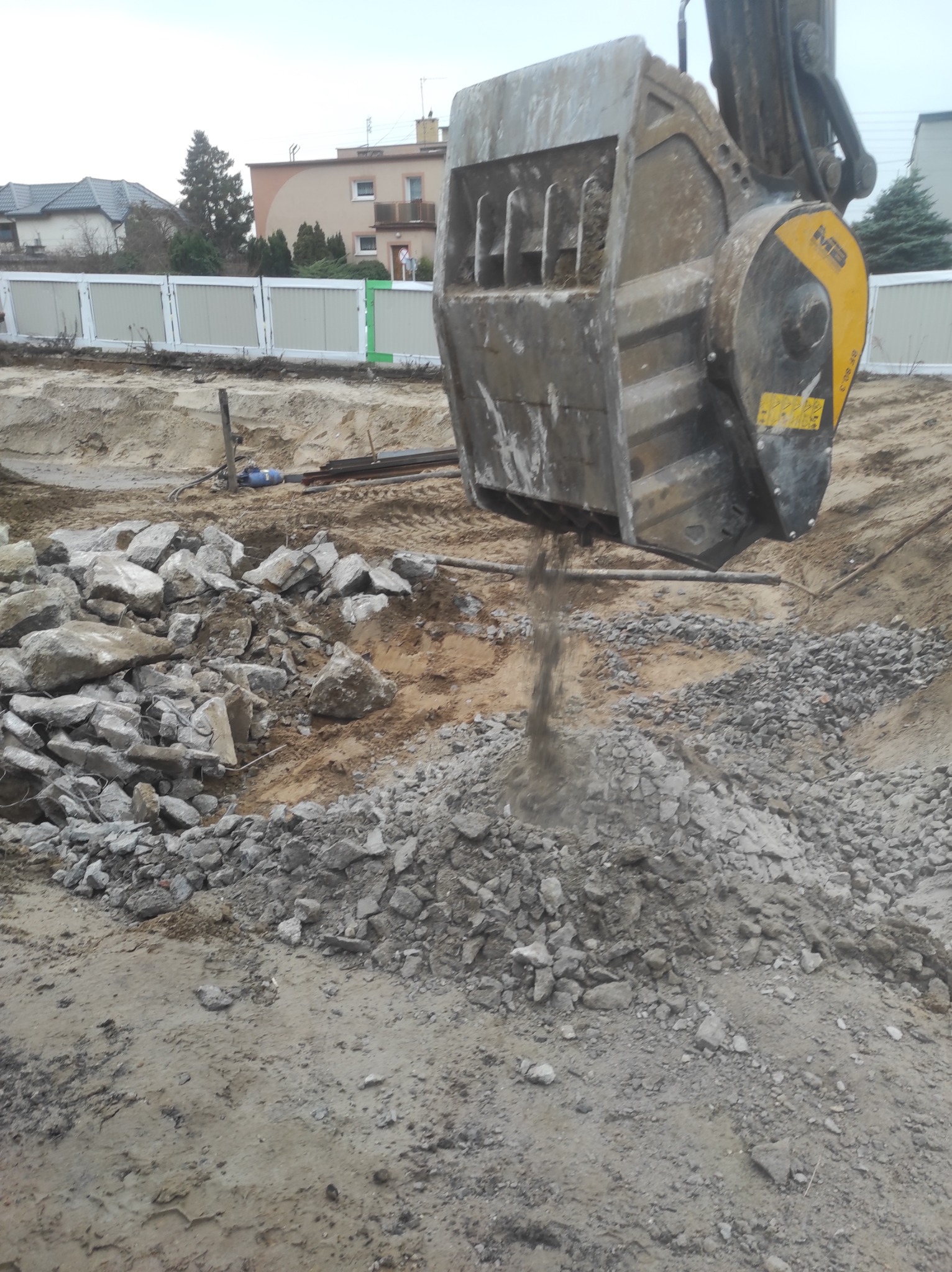 Kruszarka szczękowa zamontowana na koparce przesiewa gruz na placu budowy, w tle widoczny płot i budynki mieszkalne.