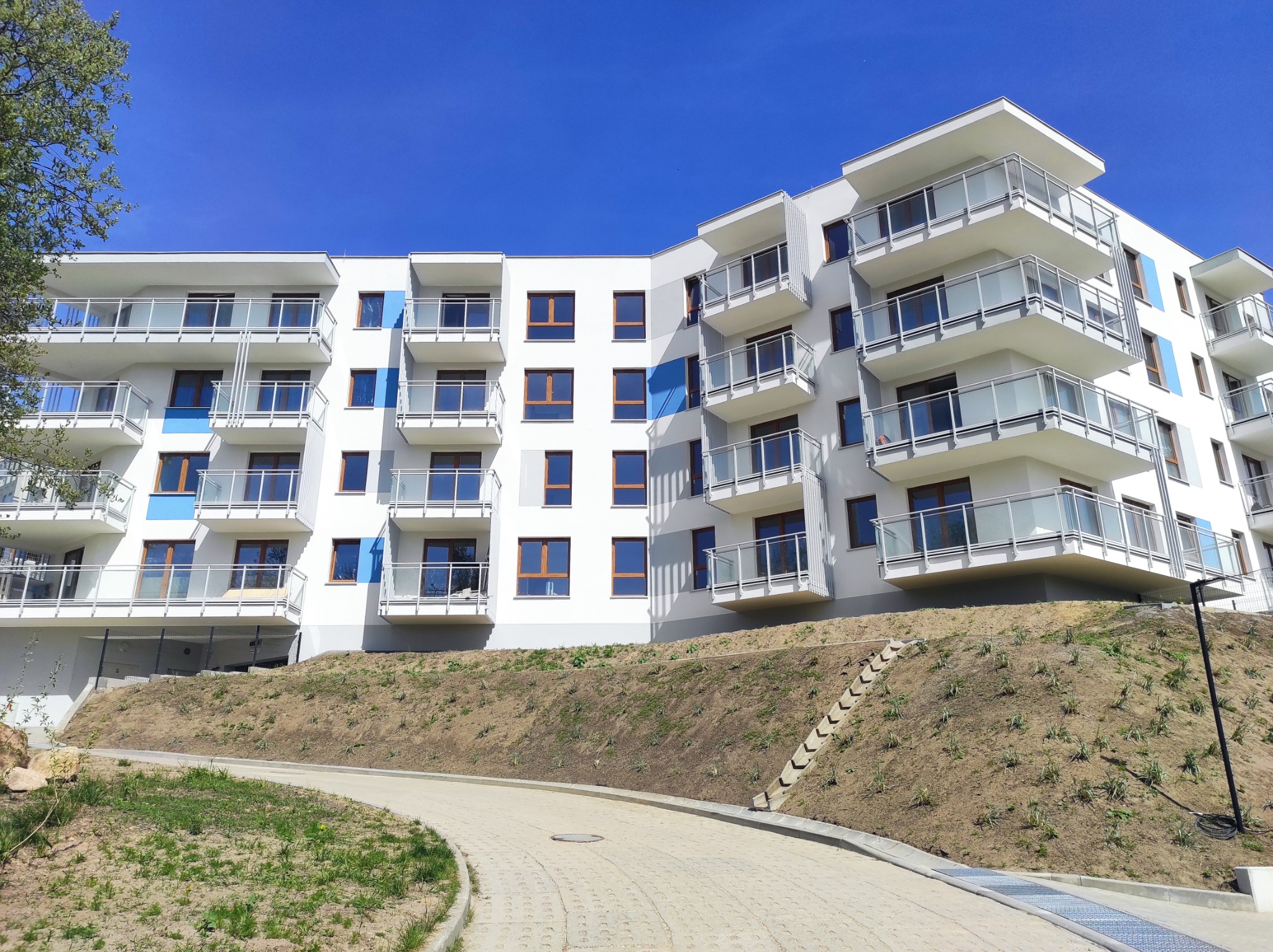 Nowoczesny, biały budynek apartamentowy z balkonami o szklanych balustradach, widoczny z poziomu chodnika prowadzącego pod górę, błękitne akcenty na elewacji.