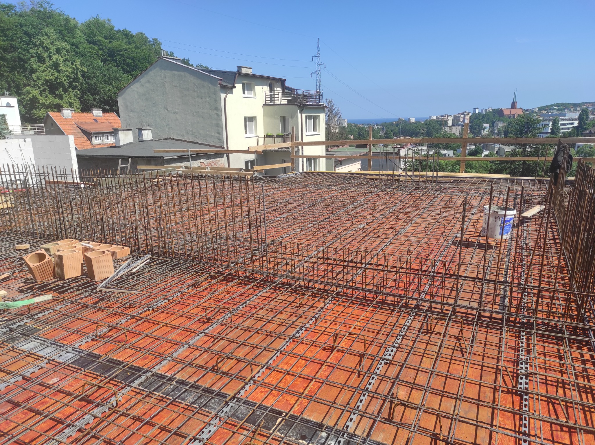 Zbrojenie stropu na pomarańczowych pustakach szalunkowych, widok z góry na konstrukcję z prętów stalowych, w tle zabudowa miejska i fragment morza.