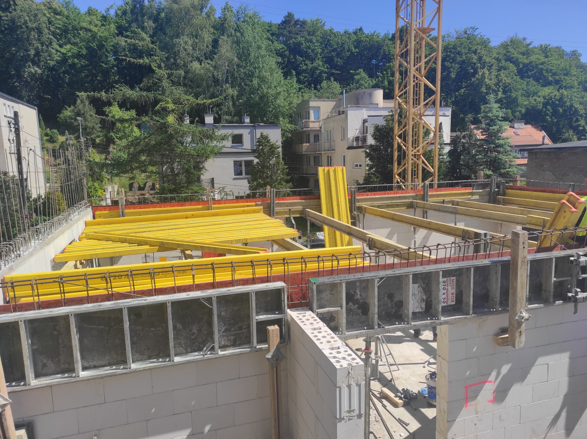 Budowa domu z białych bloczków, widoczne szalunki i żółte belki stropowe, w tle drzewa i budynki mieszkalne, częściowo widoczny żuraw budowlany.