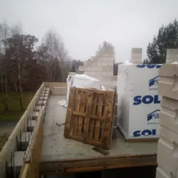 Wznoszenie ścian z bloczków betonu komórkowego na stropie budynku w trakcie budowy, widoczna paleta i materiały budowlane na tle jesiennego krajobrazu.