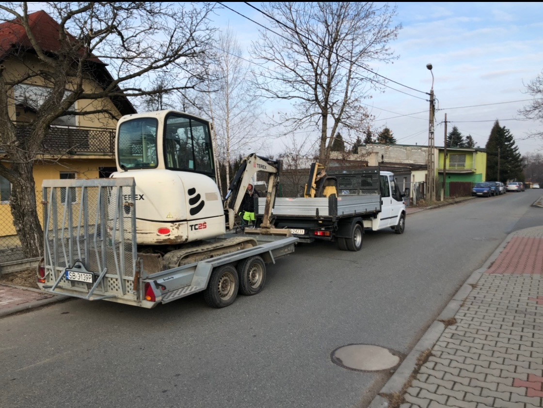 Biała mini koparka Terex TC25 na przyczepie, ciągnięta przez białego Forda Transita, na tle ulicy i zabudowań mieszkalnych.