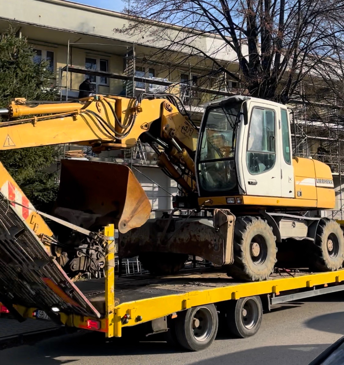 Żółto-biała koparka Liebherr na żółtej lawecie, transportowana na tle budynku w rusztowaniach.