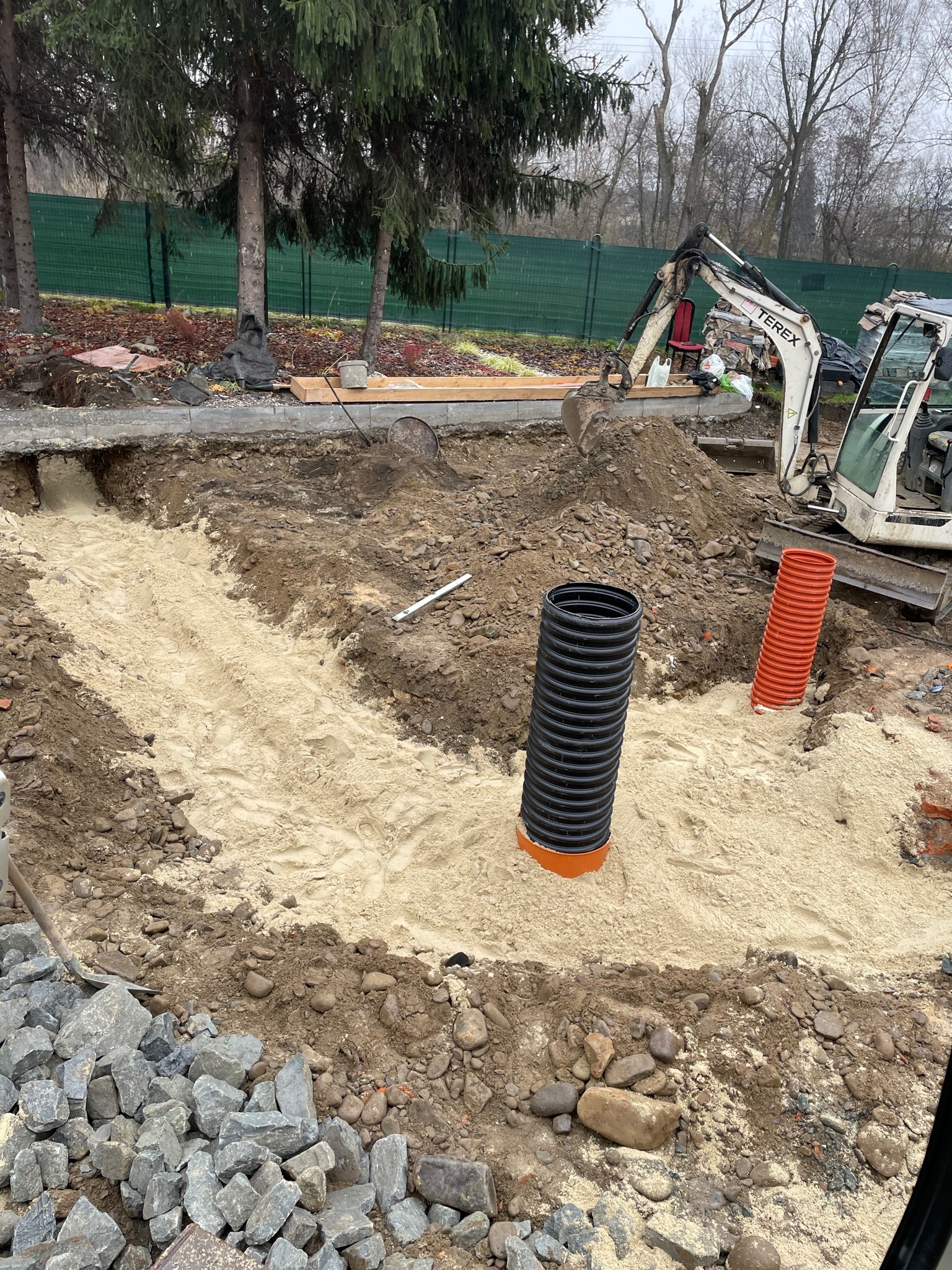 Wykop z piaskowym podsypem, widoczne rury kanalizacyjne z tworzywa sztucznego i mała koparka Terex w tle.