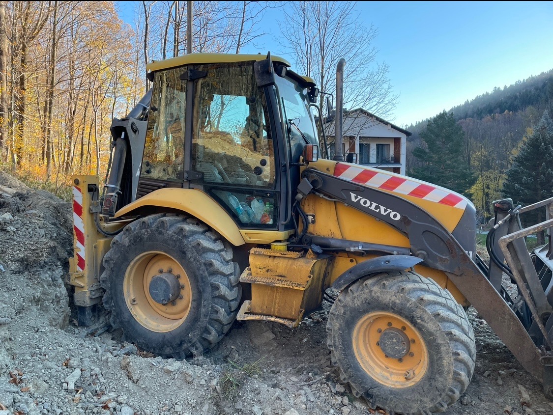 Żółta koparko-ładowarka Volvo w trakcie prac ziemnych na nierównym terenie, widoczny wykop i drzewa w tle.