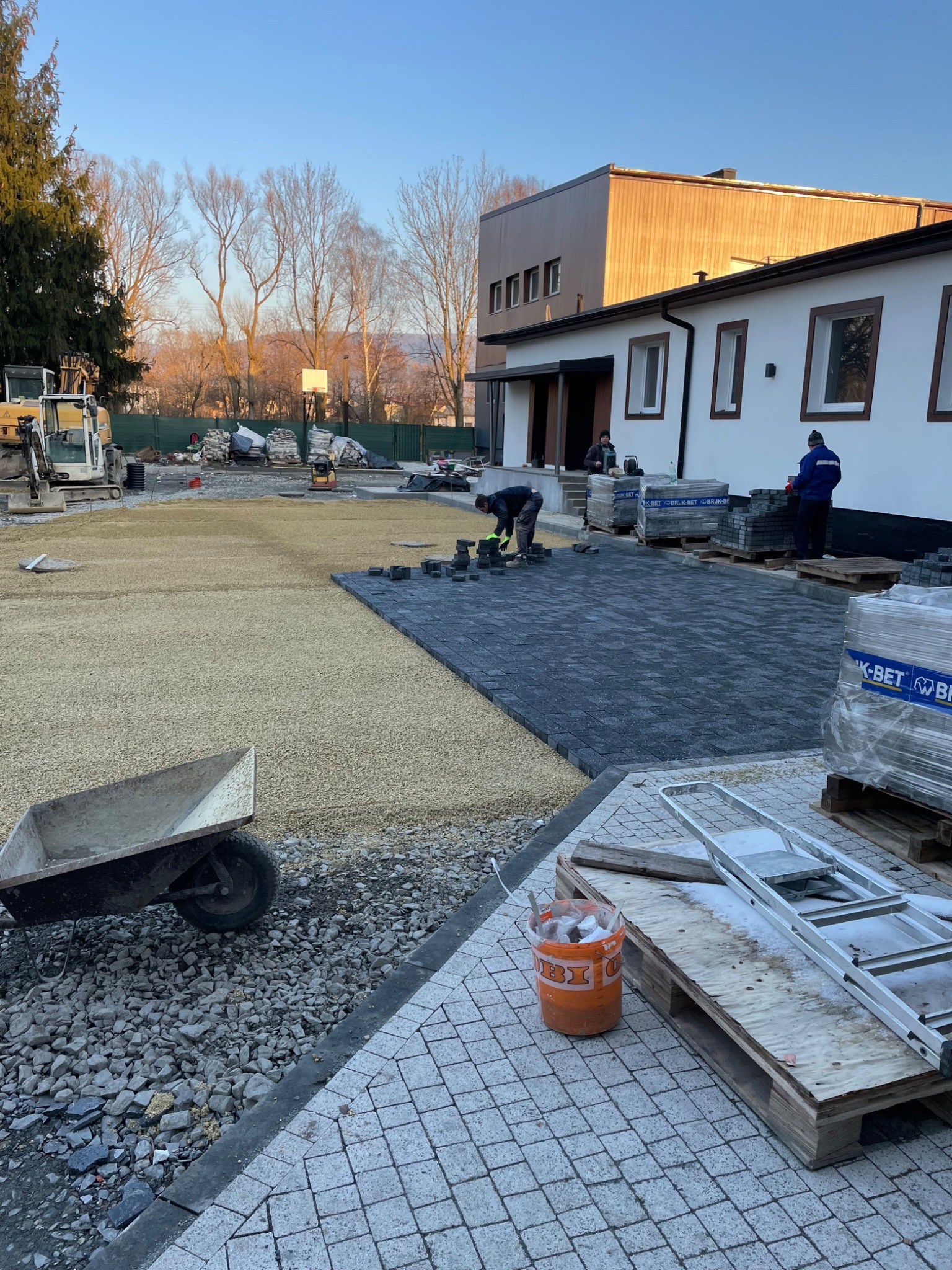 Prace brukarskie w toku: ułożona część ciemnoszarej kostki, widoczny pracownik układający kolejne elementy, przygotowane podłoże z piasku, taczka i palety z kostką w tle, budynek z białymi ścianami...