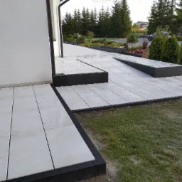 Nowoczesna posesja z jasnoszarą kostką brukową, obrzeża z ciemnej kostki, rabaty z zielenią i kwiatami, widok na dom i ogród.