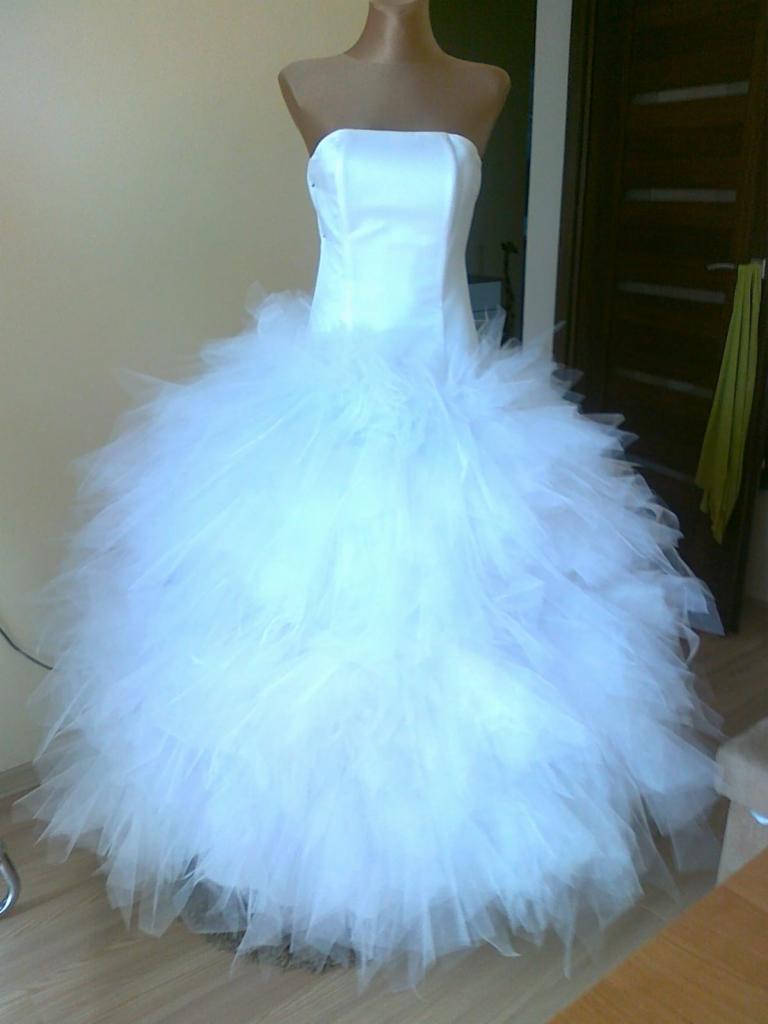 Biała suknia ślubna bez ramiączek z tiulowym dołem na manekinie, widoczne drzwi w tle.