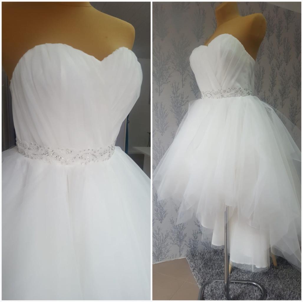 Biała sukienka z tiulu na manekinie, ozdobiona pasem z koralików, widok z przodu i z boku na tle wzorzystej tapety i dywanu.