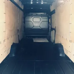 Wnętrze pustego, wyłożonego sklejką busa transportowego z czarną podłogą i oświetleniem sufitowym, przygotowanego do załadunku.