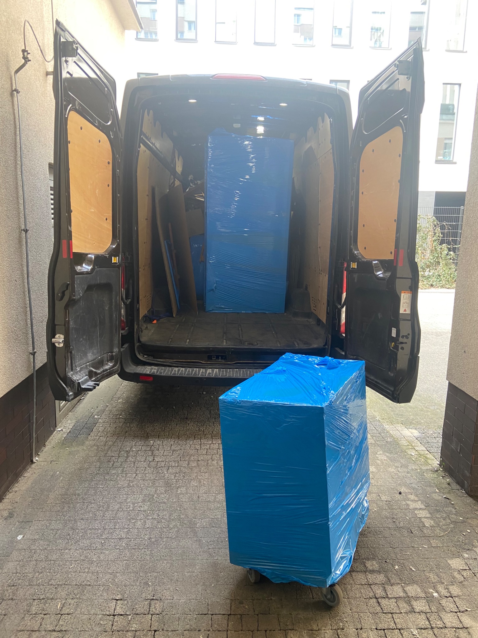 Czarny furgon z otwartymi drzwiami załadunkowymi, w środku częściowo załadowane przedmioty owinięte niebieską folią, na zewnątrz paczka na kółkach, również owinięta niebieską folią.
