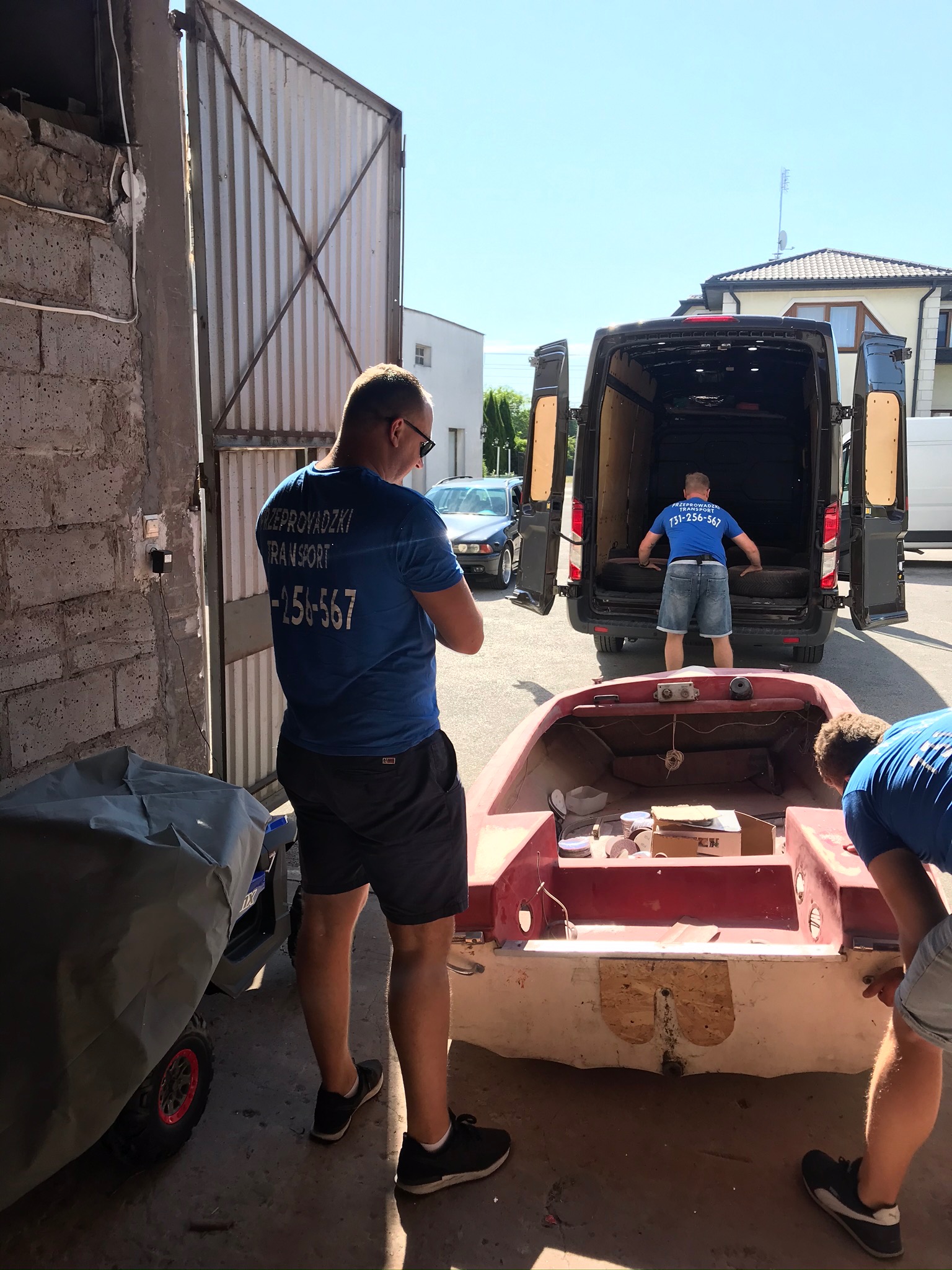 Ekipa transportowa ładuje czerwoną łódź do otwartego busa przed garażem; jeden mężczyzna w niebieskiej koszulce z napisem 'Przeprowadzki Transport' stoi i obserwuje, drugi układa opony w busie,...