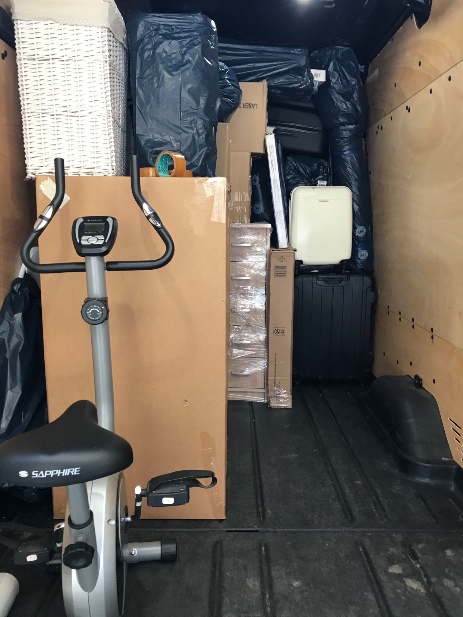 Wnętrze furgonetki załadowane do przeprowadzki: rower treningowy Sapphire, zapakowane kartony, worki i walizki zabezpieczone na czas transportu.