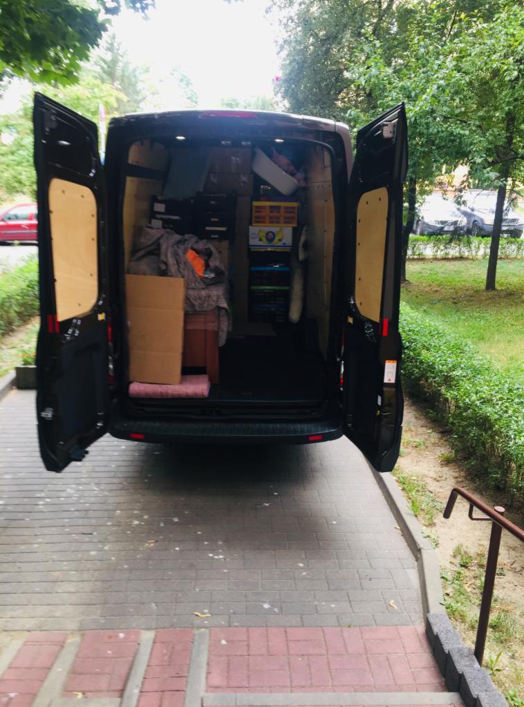 Tył czarnego busa z otwartymi drzwiami, wypełniony kartonami i przedmiotami, stojącego na chodniku z kostki brukowej, z widocznym trawnikiem i drzewami w tle.