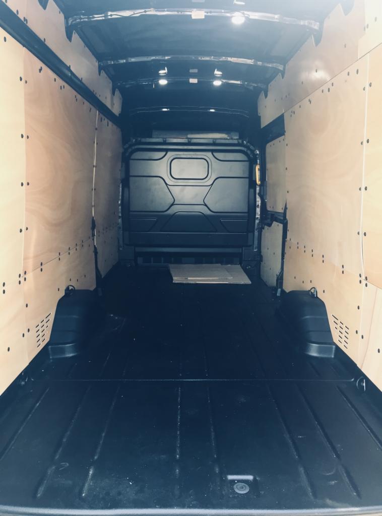 Wnętrze pustego, wyłożonego sklejką busa transportowego z czarną podłogą i oświetleniem sufitowym, przygotowanego do załadunku.