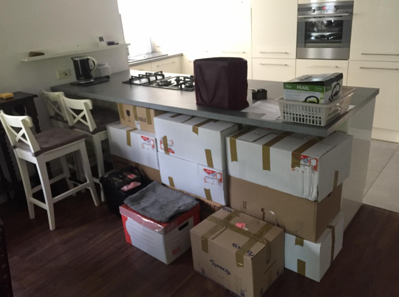 Kartonowe pudła, niektóre z naklejkami ostrzegawczymi 'fragile', przygotowane do transportu, ustawione pod blatem kuchennym w mieszkaniu w Warszawie.