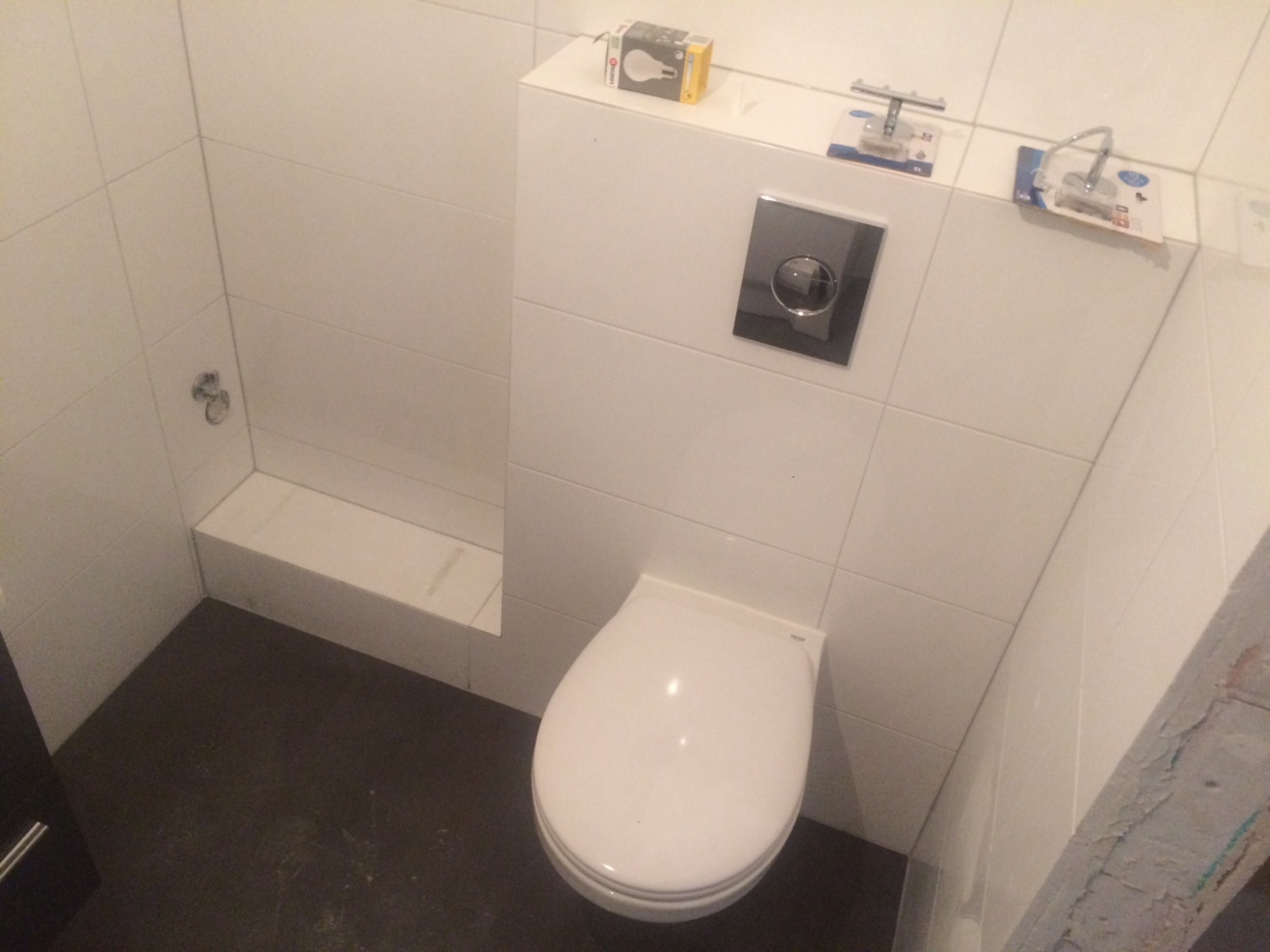 Nowoczesna toaleta podwieszana z czarnym przyciskiem spłukującym, białe płytki na ścianach i ciemna podłoga, na górze obudowy stelaża widoczne akcesoria łazienkowe w opakowaniach