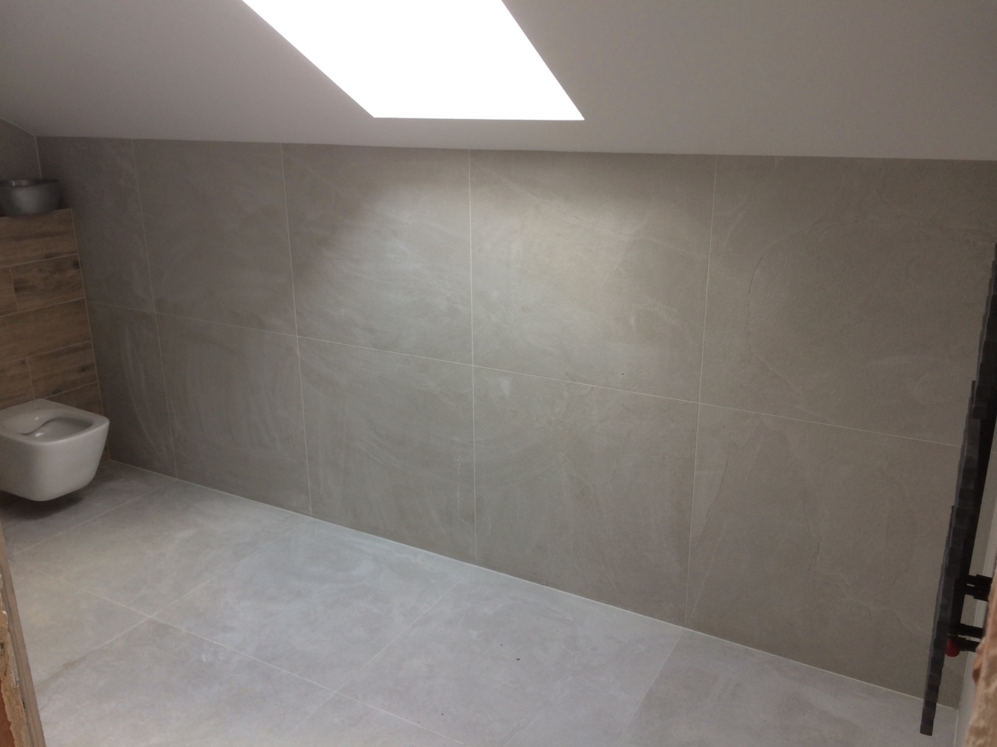 Wykończona łazienka z podwieszaną toaletą, szarymi płytkami na ścianach i podłodze oraz oświetleniem sufitowym.