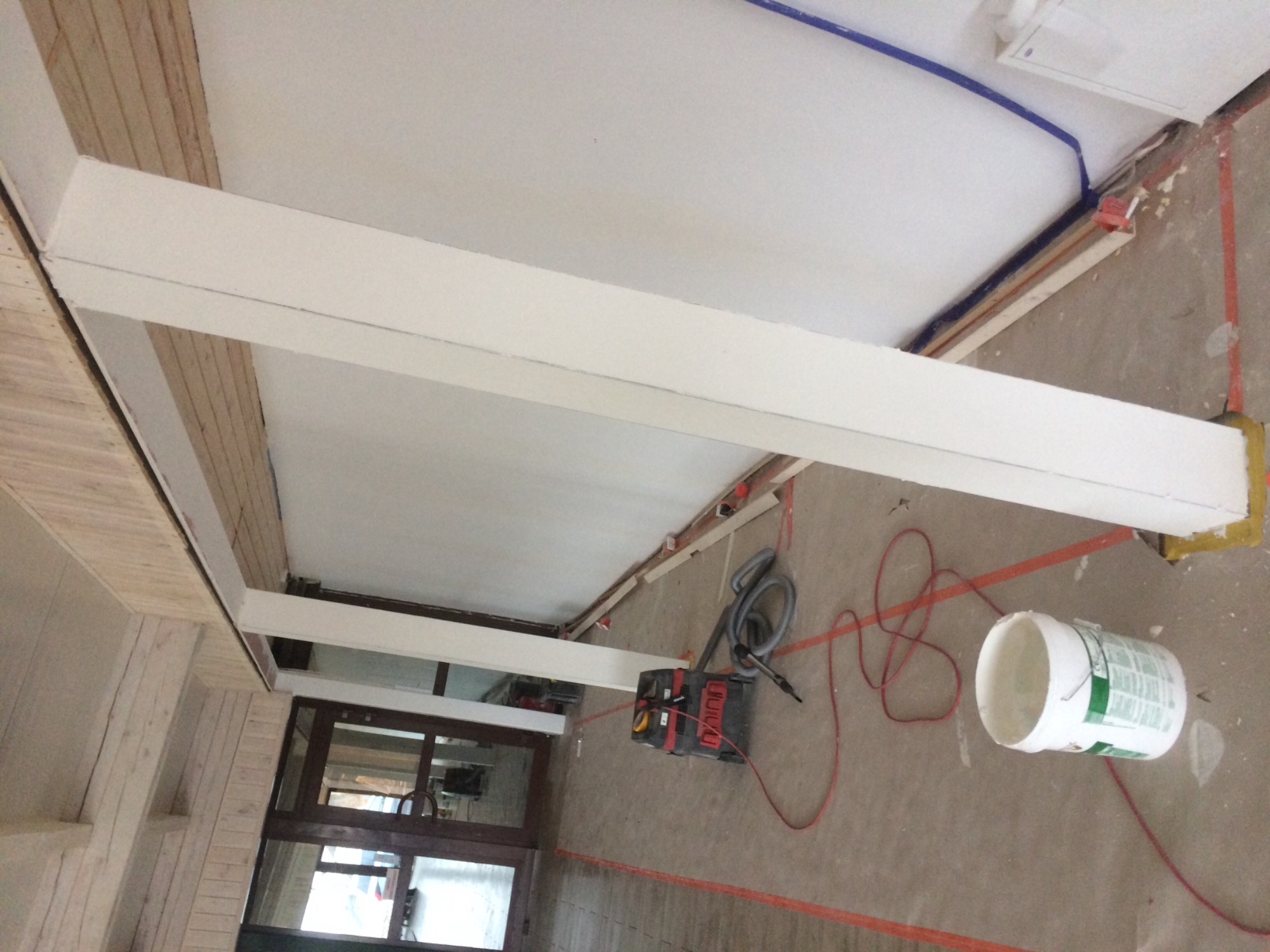 Montaż podwieszanego sufitu z białymi belkami konstrukcyjnymi, widoczny od dołu, z narzędziami i wiaderkiem na podłodze w trakcie prac.