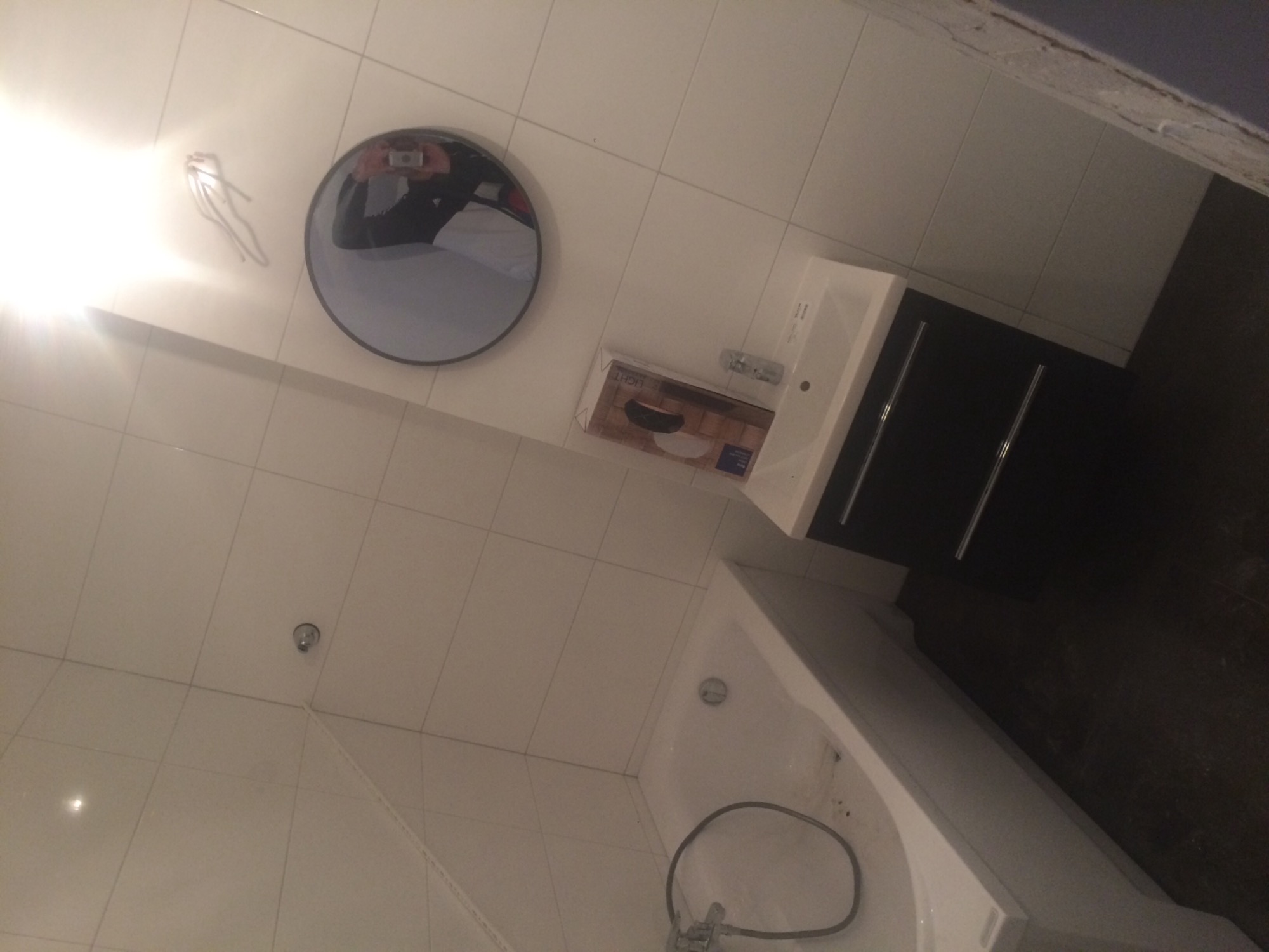 Nowa biała łazienka z umywalką z czarną szafką, okrągłym lustrem z odbiciem osoby robiącej zdjęcie, białą wanną i dozownikiem mydła w płynie.