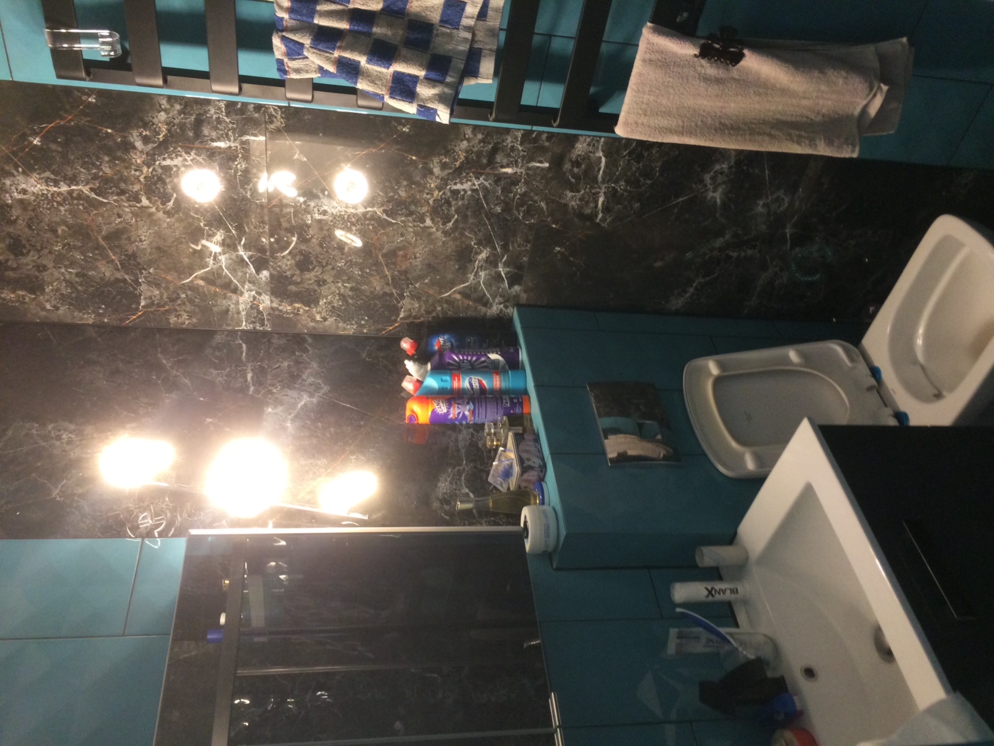 Wykończona łazienka z czarnymi marmurowymi płytkami na ścianach, turkusowymi płytkami na ścianach i podłodze, białą umywalką z czarnym blatem oraz toaletą. Na grzejniku suszarka na ręczniki...