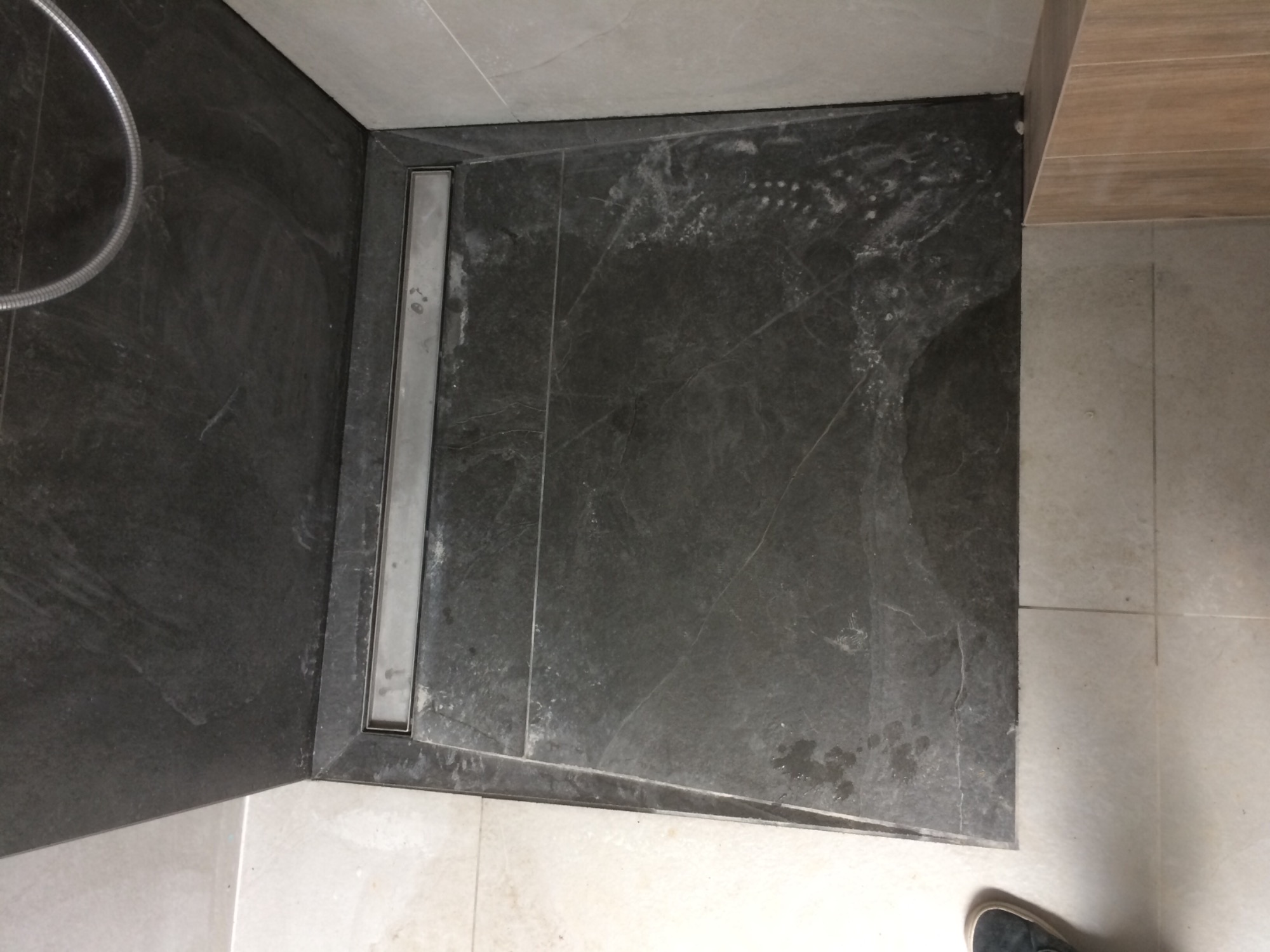 Szara, marmurkowa kabina prysznicowa z odpływem liniowym, widoczne ślady po wodzie i fragment węża prysznicowego.