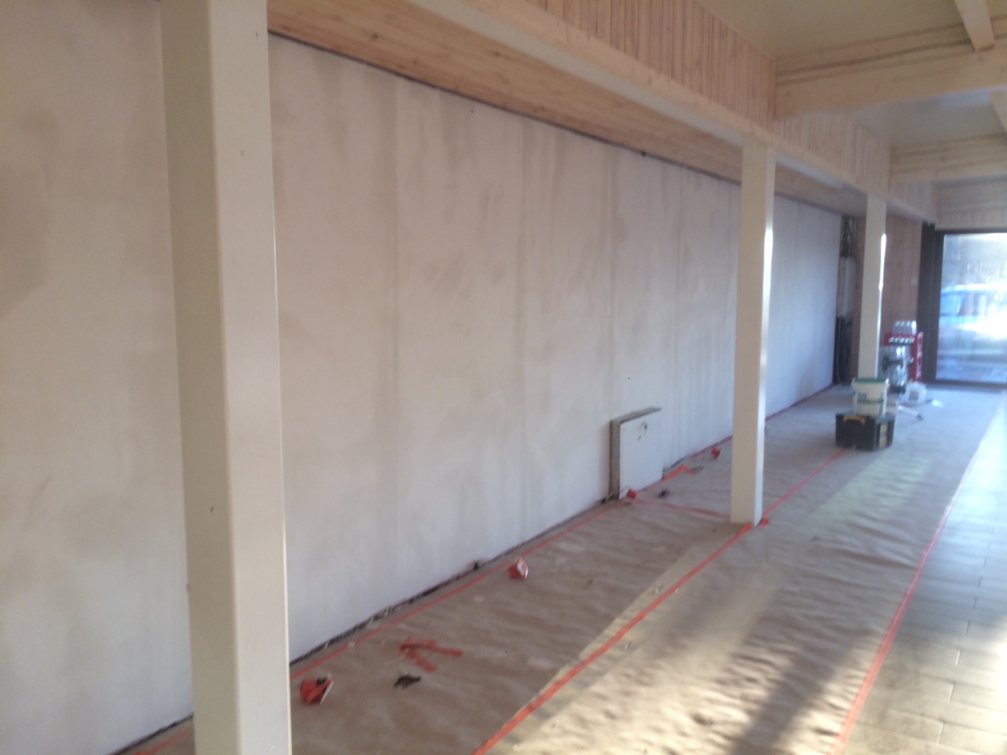 Przygotowanie do malowania ścian w pomieszczeniu z drewnianym sufitem i kolumnami, podłoga zabezpieczona folią i taśmą malarską, widoczne narzędzia malarskie.