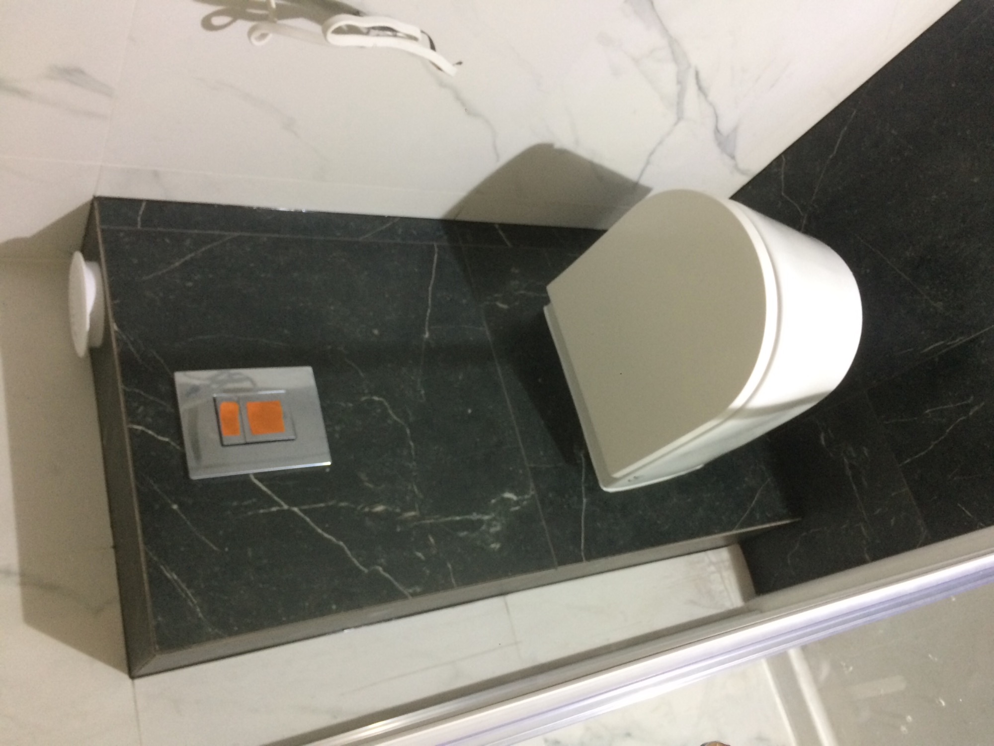 Zabudowana toaleta z czarnym blatem imitującym marmur i przyciskiem spłukiwania z pomarańczowymi akcentami, na tle białych marmurowych płytek.