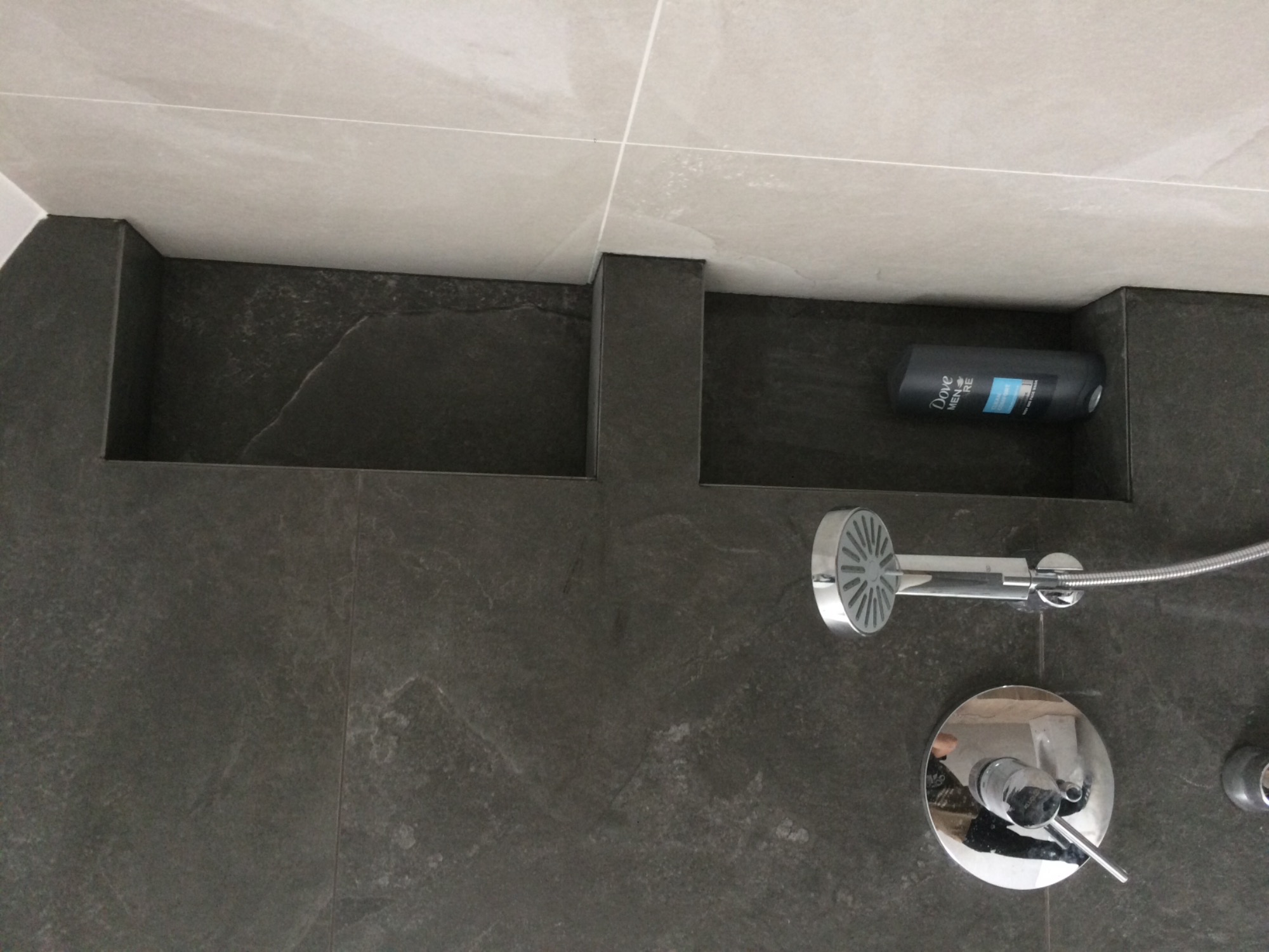 Wnęki prysznicowe wykończone ciemnym gresem, z widoczną baterią prysznicową i kosmetykiem Dove Men+Care w jednej z wnęk.