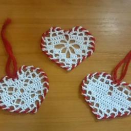 Beauty crochet - Trzy ręcznie robione, ażurowe serduszka z białej włóczki z czerwonym obramowaniem i czerwonymi sznureczkami do zawieszenia, leżące na drewnianym blacie.