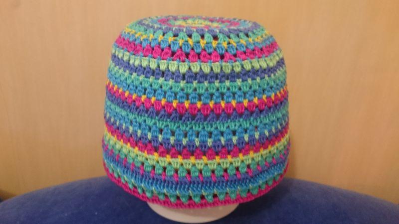 Czapka dziergana na szydełku z włóczki w kolorach: różowym, żółtym, zielonym i niebieskim, leżąca na niebieskim materiale, na beżowym tle.