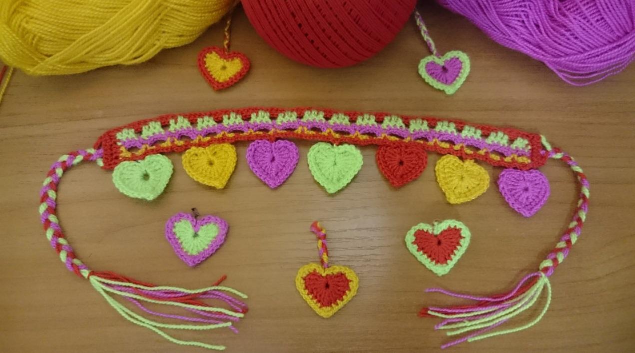 Ręcznie robiona, kolorowa opaska na włosy z szydełkowymi serduszkami w odcieniach zieleni, fioletu, czerwieni i żółci, leżąca na drewnianym stole obok kłębków włóczki w tych samych kolorach. Opaska...